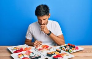 Young man sitting at a table with sushi selection feeling unwell after eating.